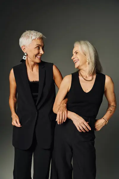 Laughing senior women in black trendy attire looking at each other on grey, lifelong friends — Stock Photo