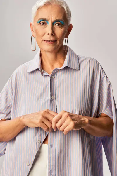 Модна старша жінка з коротким срібним волоссям і сміливим макіяжем у синій смугастій сорочці на сірому — стокове фото
