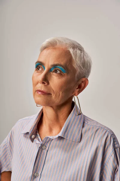 Stylisches, verträumtes Seniormodel mit kurzen silbernen Haaren und blauem Eyeliner, das auf Grau wegschaut, Porträt — Stockfoto