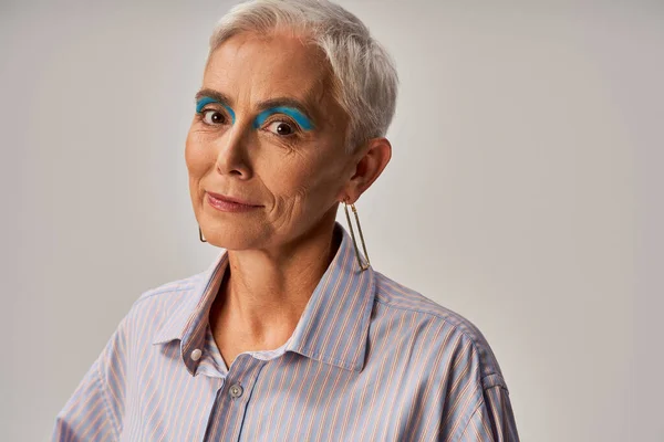 Portrait of elegant mature lady with short silver hair and bold makeup smiling at camera on grey — Stock Photo