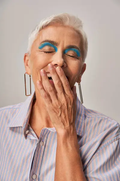 Aufgeregte reife Frau mit kühnem Make-up, die mit geschlossenen Augen lacht und den Mund mit der Hand auf grau abdeckt — Stockfoto