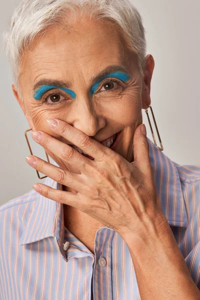 Senhora madura alegre com olhos azuis rindo da câmera e cobrindo a boca com a mão no cinza — Fotografia de Stock
