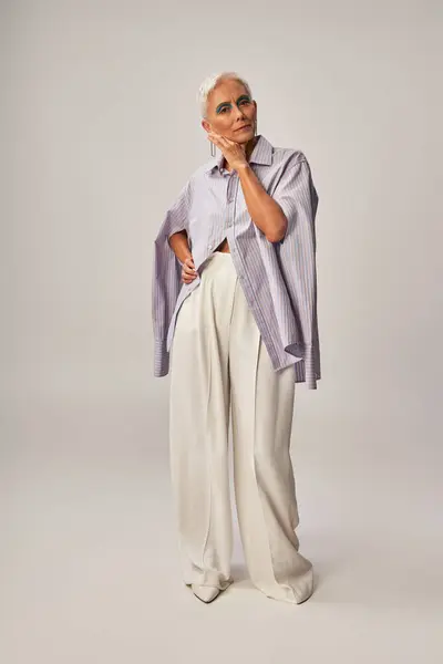 Trendy aging, full length of senior woman in blue striped shirt and white pants standing on grey — Stock Photo