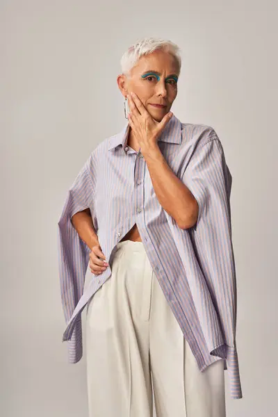 Mujer mayor en camisa de rayas azules y pantalones blancos tocando la cara y mirando a la cámara en gris - foto de stock