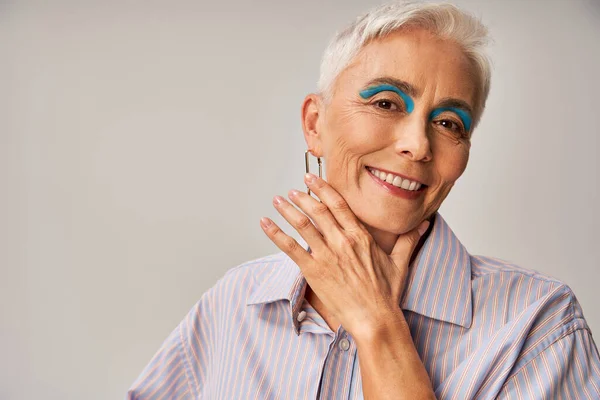 Cheerful mature lady with blue eyeliner and short silver hair looking at camera on grey backdrop — Stock Photo