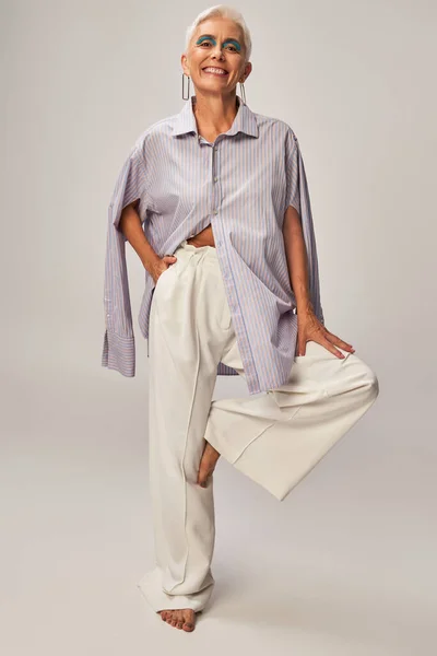 Joyful and barefoot mature lady in blue striped shirt posing on one leg with hand in pocket on grey — Stock Photo