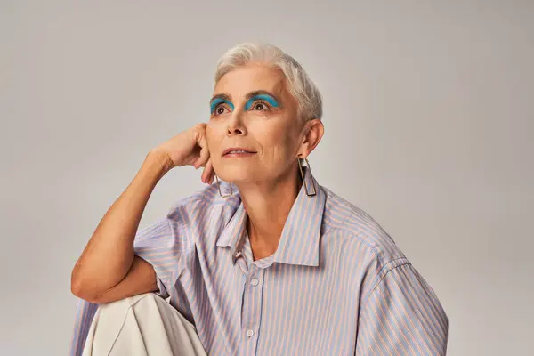 Pensive and stylish senior woman in blue striped shirt and bold makeup looking away on grey — Stock Photo