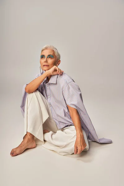 Barefoot and dreamy senior model in blue striped shirt and pants sitting and looking away on grey — Stock Photo