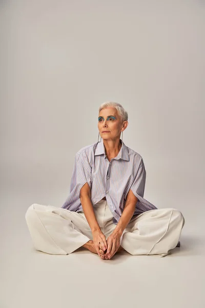 Pensive and barefoot mature lady in stylish casual clothes looking away while sitting on grey — Stock Photo