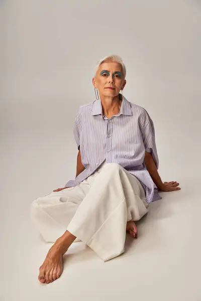 Descalza y confiada señora mayor en traje casual de moda mirando a la cámara mientras está sentado en gris - foto de stock
