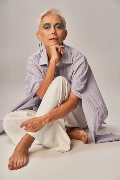 Модная стареющая, босиком зрелая женщина в голубой полосатой рубашке сидит и смотрит в камеру на сером — стоковое фото