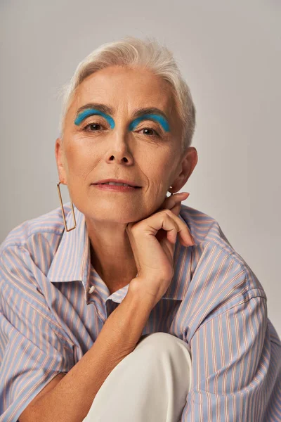 Mature lady with bold makeup posing in blue striped shirt and smiling at camera on grey — Stock Photo