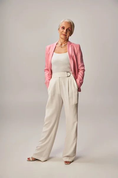 Повна довжина старшої моделі в рожевому блістері, що стоїть руками в кишенях білих штанів на сірому — стокове фото