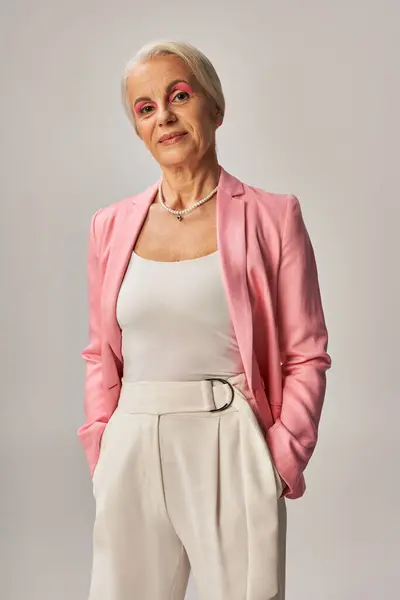 Smiling mature woman in pink blazer posing with hands in pockets of white pants on grey backdrop — Stock Photo