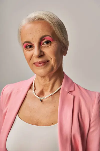 Portrait of elegant senior lady in pink blazer and pearl necklace smiling at camera on grey — Stock Photo