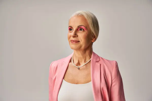Dreamy senior woman in pink blazer and pearl necklace smiling and looking away on grey, elegance — Stock Photo