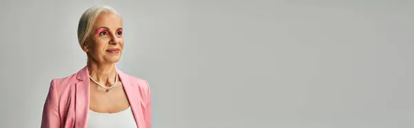 Мечтательная и стильная зрелая леди в розовом блейзере и жемчужном ожерелье глядя в сторону на серый, баннер — стоковое фото