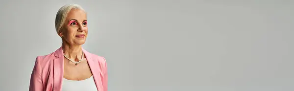 Dreamy and stylish mature lady in pink blazer and pearl necklace looking away on grey, banner — Stock Photo