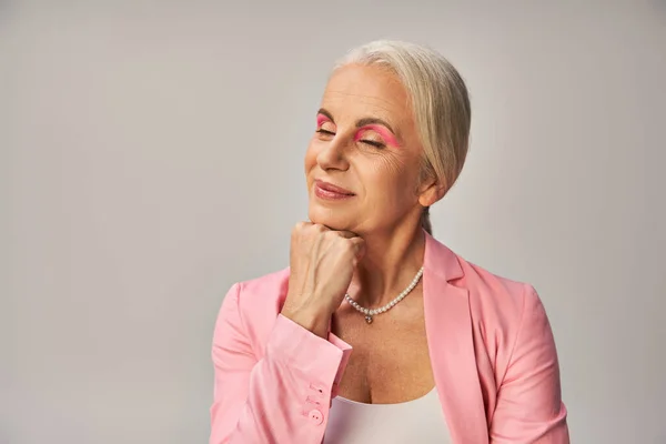 Мрійлива і усміхнена старша жінка в рожевому блістері тримає руку близько і позує із закритими очима на сірому — стокове фото