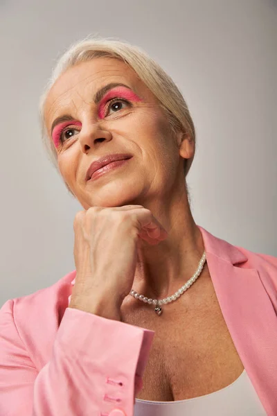 Portrait of dreamy senior model in pink blazer holding hand near chin and looking away on grey — Stock Photo