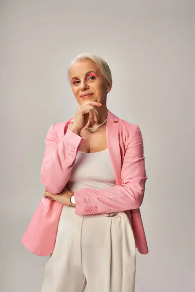 Положительный старший женщина в розовом блейзере стоя с рукой возле подбородка и глядя на камеру на сером — стоковое фото