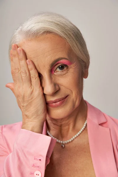 Portrait of elegant mature lady in pearl necklace and pink blazer obscuring face with hand on grey — Stock Photo