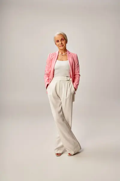 Full length of mature lady in pink blazer posing with hands in pockets of white pants on grey — Stock Photo