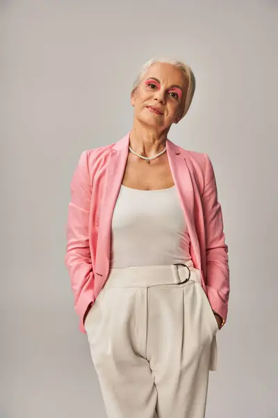 Señora madura de moda en blazer rosa de pie con las manos en bolsillos de pantalones blancos en gris, elegancia - foto de stock