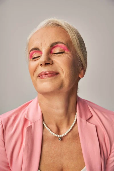 Porträt einer lächelnden Seniorin in rosa Blazer und Perlenkette, die mit geschlossenen Augen auf grau posiert — Stockfoto