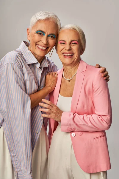 Fröhliche Seniorinnen in stilvoller blauer und pinkfarbener Kleidung umarmen und lächeln in die Kamera auf grau — Stockfoto