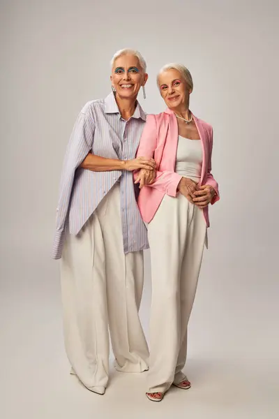 Mulheres seniores na moda e alegres em traje casual elegante sorrindo para a câmera em cinza, comprimento total — Fotografia de Stock