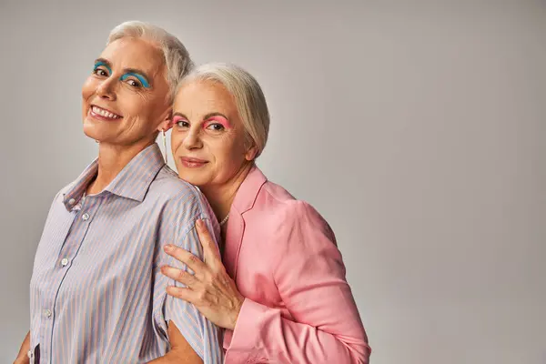 Encantadas amigas mayores en ropa azul y rosa sonriendo a la cámara en gris, positividad de edad - foto de stock