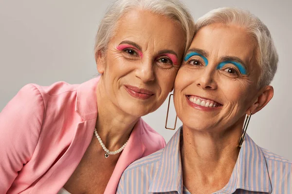 Allegre signore anziane in abiti blu e rosa e trucco in posa su grigio, moda e amicizia — Foto stock