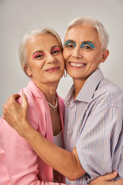 Mujer mayor feliz con delineador de ojos azul abrazando elegante amiga femenina en gris, envejecimiento de moda - foto de stock