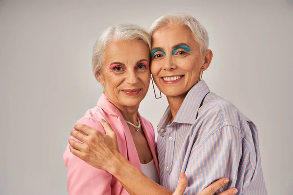 Fashionable senior female friends in blue and pink clothes embracing and smiling at camera on grey — Stock Photo