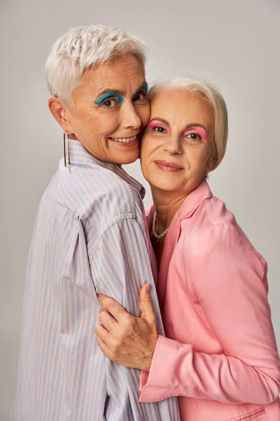 Gioiose fashioniste senior in abiti blu e rosa che abbracciano e guardano la fotocamera su sfondo grigio — Foto stock