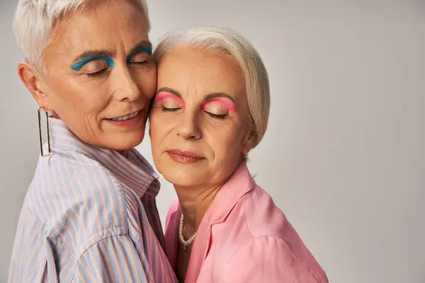 Portrait of mature female friends with silver hair and makeup standing with closed eyes on grey — Stock Photo