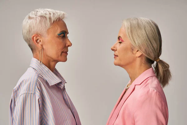 Side view of fashionable senior ladies in blue and pink attire standing on grey, vanity fair concept — Stock Photo