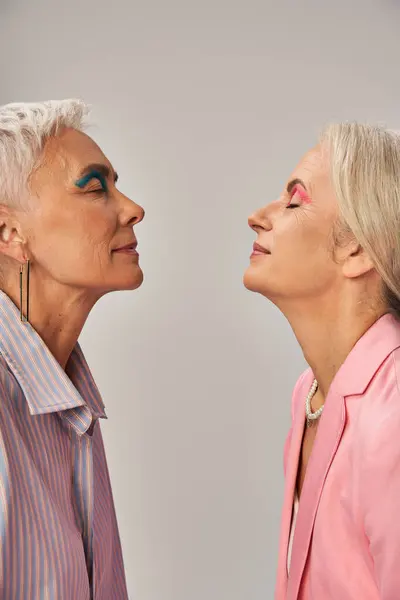 Élégantes amies aînées en vêtements bleus et roses debout avec les yeux fermés sur le gris, vue latérale — Photo de stock