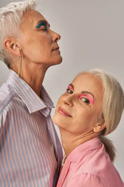 Compiaciuta donna anziana guardando la fotocamera vicino amico femminile alla moda con trucco blu su sfondo grigio — Foto stock