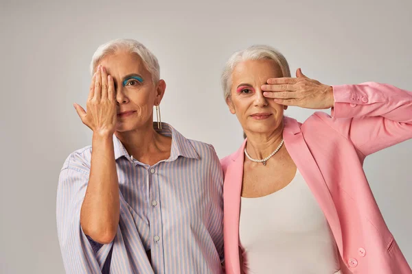 Trendy mature models in blue and pink wear obscuring faces with hands and looking at camera on grey — Stock Photo
