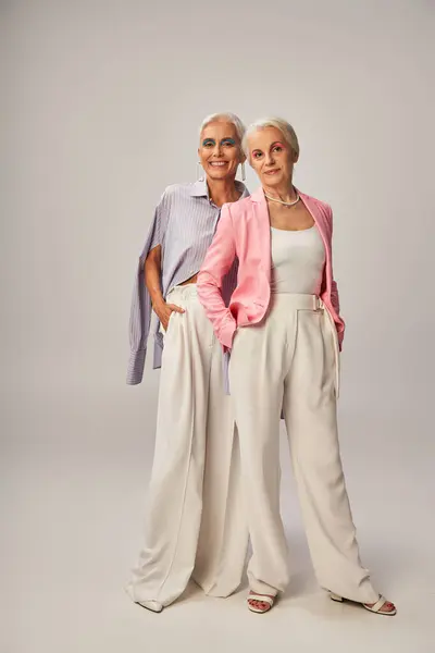 Full length of happy senior female friends in trendy clothes standing with hands in pockets on grey — Stock Photo