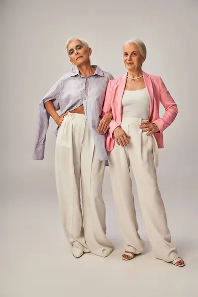 In voller Länge lächelnde Seniorinnen in stylischer Kleidung posieren mit den Händen auf den Hüften auf grau — Stockfoto