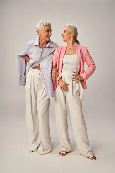 Fashionable senior female friends standing with hands on hips and smiling at each other on grey — Stock Photo