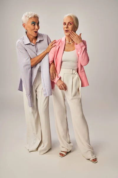Amazed senior woman looking at stylish female friend touching face on grey, ageless beauty — Stock Photo