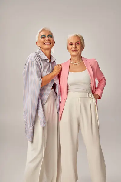 Alegres señoras mayores en atuendo casual de moda mirando a la cámara en gris, concepto de envejecimiento feliz - foto de stock