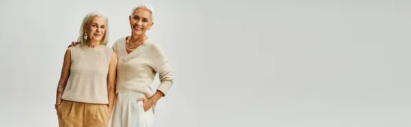 Gioiose signore mature in abiti alla moda pastello in piedi con le mani in tasca su grigio, banner — Foto stock