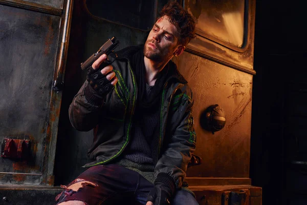 Uomo non rasato in abiti consumati guardando pistola vicino alla carrozza della metropolitana arrugginita, sopravvivenza post-disastro — Foto stock