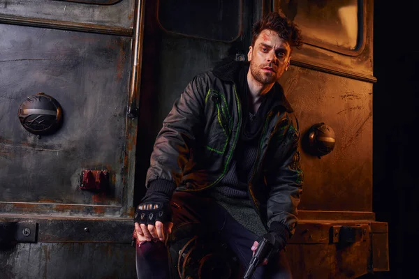 Injured man in worn jacket holding gun and looking at camera near rusty carriage in dark subway — Stock Photo