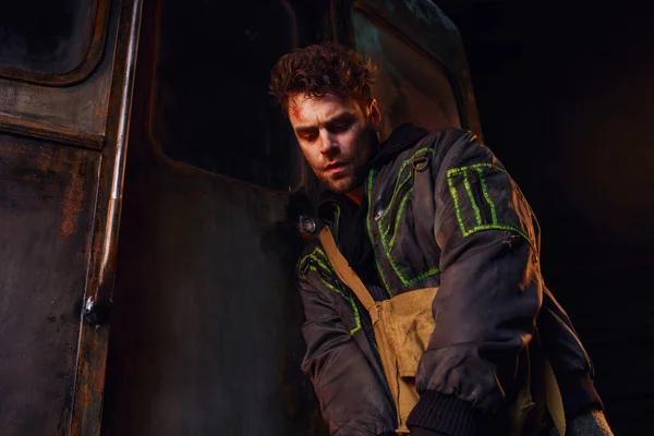 Небритый раненый мужчина в ношеной куртке возле ржавой кареты в темном метро, последствия катастрофы — стоковое фото